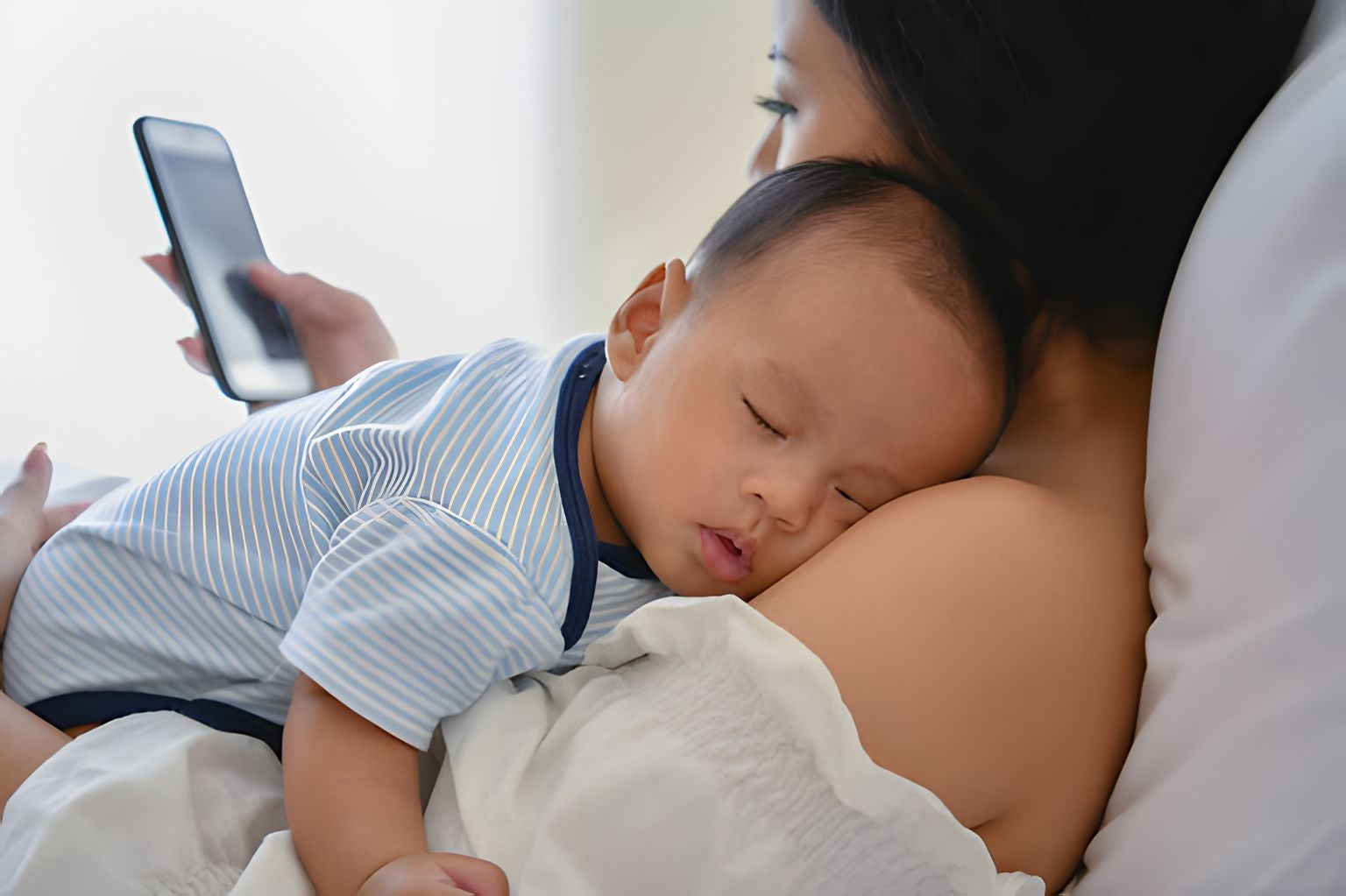 New mom laying in bed, holding her newborn and her phone.