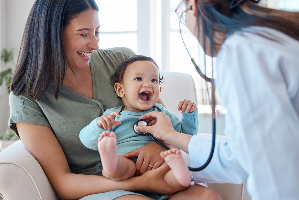 Young mom holding her baby while a pediatrician checks him with her stethoscope.